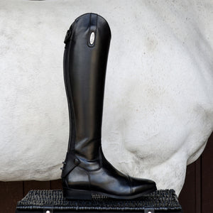 A single black leather equestrian riding boot with a pointed toe and side buckle detail, standing upright on a woven black platform in front of a white horse.