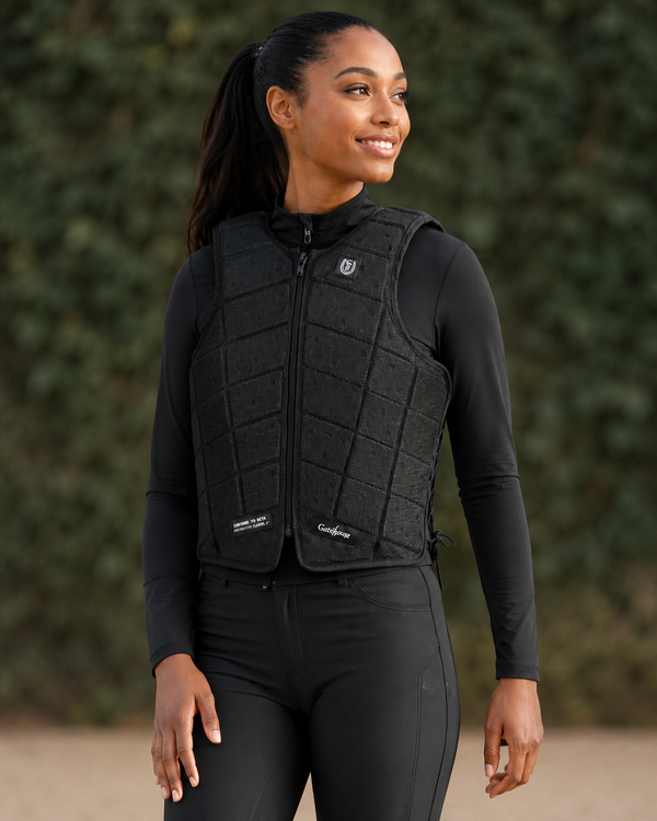 A woman wearing a black quilted equestrian protective vest over a black long-sleeve shirt and black riding pants, standing outdoors against a blurred green hedge background.