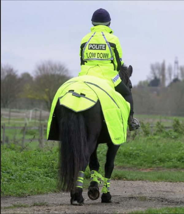 A police officer on horseback wearing a bright high-visibility neon yellow riding jacket and matching leg wraps, riding a black horse along a dirt path outdoors.